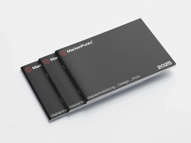 Drei schwarze Broschüren für das Jahr 2025 mit der Aufschrift „MarkenPunkt“ liegen gestapelt auf einer weißen Oberfläche. Auf den Covern ist der Text „Webentwicklung – Design – Print“ zu sehen.